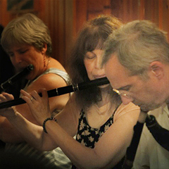 Three people play instruments in an Irish session.  Lesl Harker is in the center, playing a black wooden keyless flute. To her left is a man playing uilleann pipes; to her right is a woman also playing a wooden flute.  They all appear to be in their 60s or 70s, and the women are dressed for summer.